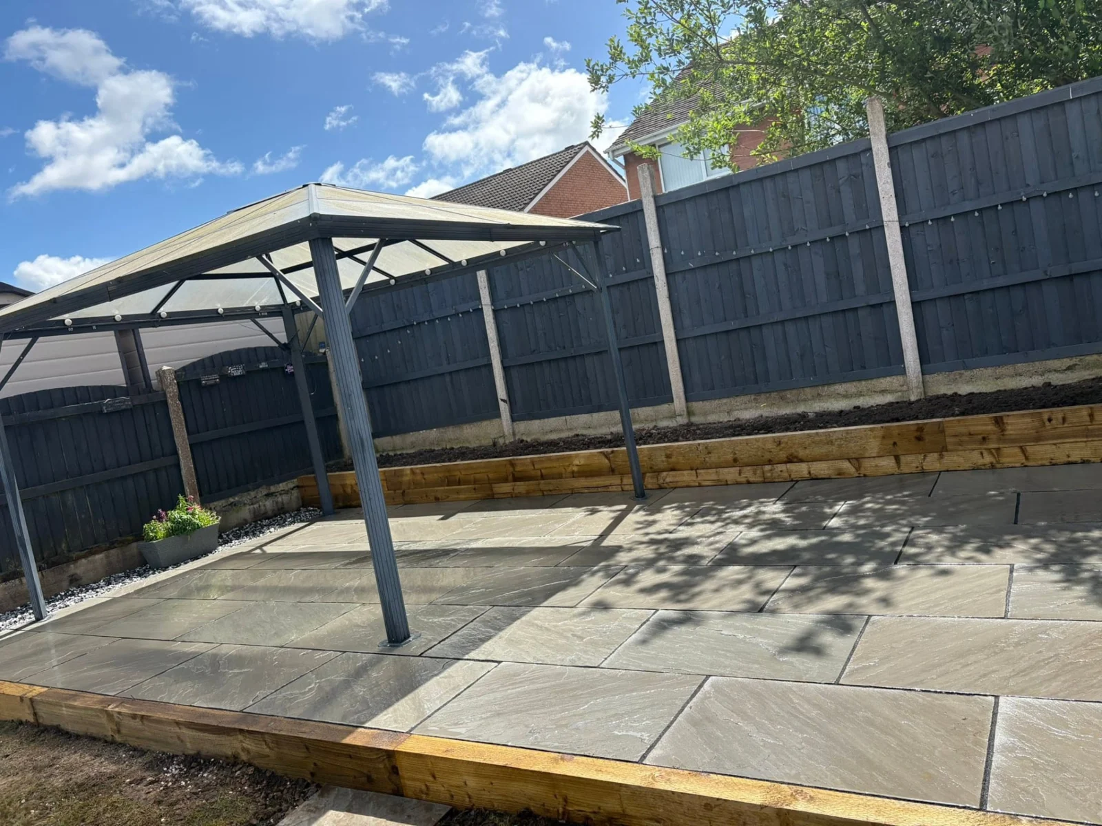 Contemporary sandstone patio with canopy, dark fencing and timber sleeper edging